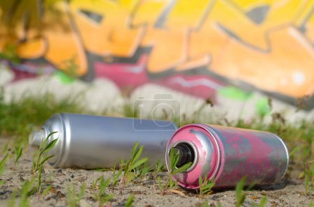 Several used spray cans with paint and caps for spraying paint under pressure on grass near the painted wall in colored graffiti drawings