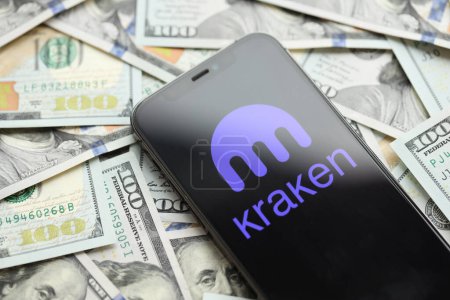 Foto de KYIV, UCRANIA - MARZO 15, 2024 Kraken logo on iPhone display screen with many hundred dollar bills. Criptomoneda portal de cambio - Imagen libre de derechos