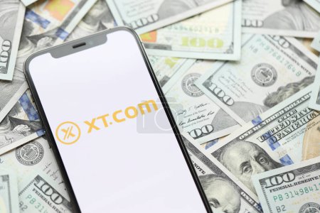 Photo for KYIV, UKRAINE - MARCH 15, 2024 XTcom logo on iPhone display screen with many hundred dollar bills. Cryptocurrency exchange portal - Royalty Free Image