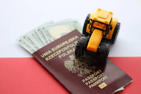 Téléchargez les photos : Passeport polonais rouge et tracteur jaune sur l'argent américain et drapeau rouge et blanc lisse de la Pologne gros plan - en image libre de droit