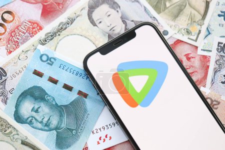 Téléchargez les photos : KYIV, UKRAINE - 1 AVRIL 2024 Tencent Video Icône WeTV sur l'écran du smartphone sur de nombreuses factures d'argent asiatiques. Écran iPhone avec logo app avec yens japonais et billets de banque yuans chinois - en image libre de droit