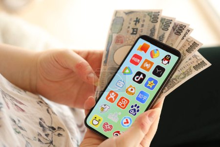 Téléchargez les photos : KYIV, UKRAINE - 1 AVRIL 2024 icône des applications asiatiques sur l'écran du smartphone et de l'argent à la main des femmes. Écran iPhone avec logo de l'application et billets d'argent en yen japonais dans les mains des femmes fermer - en image libre de droit
