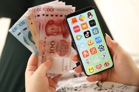 Téléchargez les photos : KYIV, UKRAINE - 1 AVRIL 2024 icône des applications asiatiques sur l'écran du smartphone et de l'argent à la main des femmes. Écran iPhone avec logo app et billets d'argent yuan chinois dans les mains des femmes fermer - en image libre de droit
