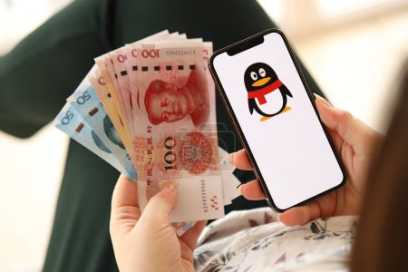Téléchargez les photos : KYIV, UKRAINE - 1 AVRIL 2024 Tencent icône QQ sur l'écran du smartphone et de l'argent dans la main des femmes. Écran iPhone avec logo app et billets d'argent yuan chinois dans les mains des femmes fermer - en image libre de droit