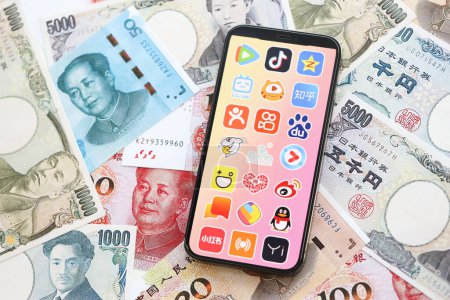 Téléchargez les photos : KYIV, UKRAINE - 1 AVRIL 2024 icône des applications asiatiques sur l'écran du smartphone sur de nombreuses factures d'argent asiatiques. Écran iPhone avec logo app avec yens japonais et billets de banque yuans chinois - en image libre de droit