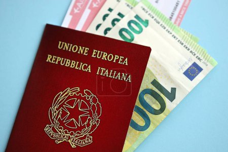 Pasaporte italiano con dinero y billetes de avión en fondo azul de cerca. Concepto de turismo y viajes