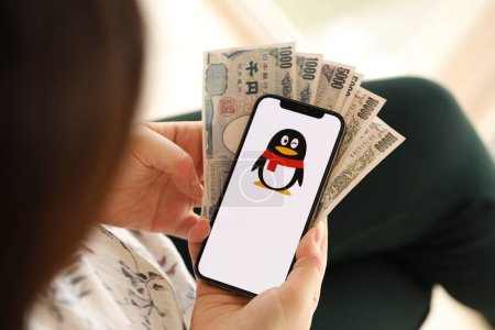 Téléchargez les photos : KYIV, UKRAINE - 1 AVRIL 2024 Tencent icône QQ sur l'écran du smartphone et de l'argent dans la main des femmes. Écran iPhone avec logo de l'application et billets d'argent en yen japonais dans les mains des femmes fermer - en image libre de droit