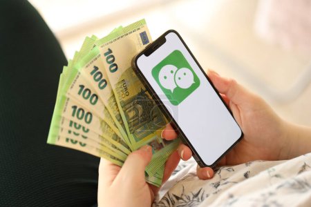 Photo for KYIV, UKRAINE - APRIL 1, 2024 Wechat or Weixin icon on smartphone screen and money in female hand. iPhone display with app logo and hundred euro bills in girls hands close up - Royalty Free Image