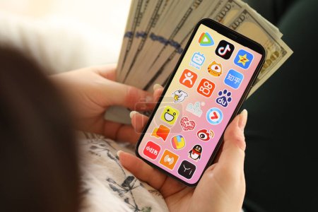 Téléchargez les photos : KYIV, UKRAINE - 1 AVRIL 2024 icône des applications asiatiques sur l'écran du smartphone et de l'argent à la main des femmes. Écran iPhone avec logo de l'application et billets de cent dollars dans les mains des filles fermer - en image libre de droit