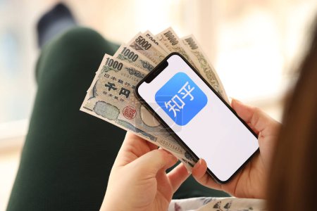 Téléchargez les photos : KYIV, UKRAINE - 1er AVRIL 2024 icône Zhihu sur l'écran du smartphone et de l'argent à la main féminine. Écran iPhone avec logo de l'application et billets d'argent en yen japonais dans les mains des femmes fermer - en image libre de droit