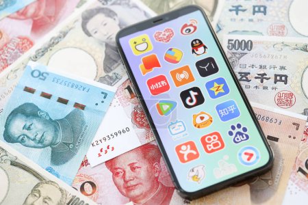 Téléchargez les photos : KYIV, UKRAINE - 1 AVRIL 2024 icône des applications asiatiques sur l'écran du smartphone sur de nombreuses factures d'argent asiatiques. Écran iPhone avec logo app avec yens japonais et billets de banque yuans chinois - en image libre de droit