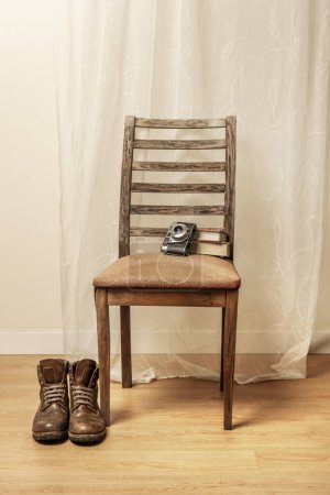 Foto de Botas de senderismo de cuero marrón que descansan junto a una silla de madera demacrada junto a una vieja cámara que descansa sobre el respaldo - Imagen libre de derechos