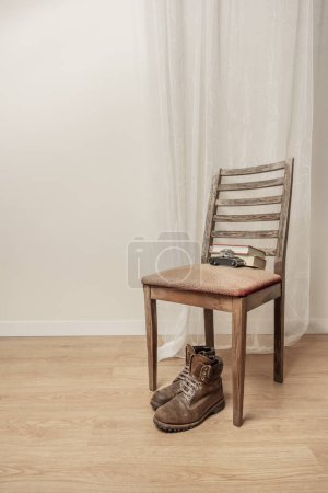 Foto de Botas de montañismo de cuero marrón que descansan bajo el asiento de una silla de madera demacrada junto a una vieja cámara con algunos libros y una sutil cortina blanca detrás. - Imagen libre de derechos