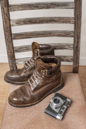 Foto de Un par de botas de cuero marrón para hombre con triple costura en una silla vieja junto a una cámara fotográfica vintage en una habitación con suelos de parquet marrón y paredes blancas - Imagen libre de derechos