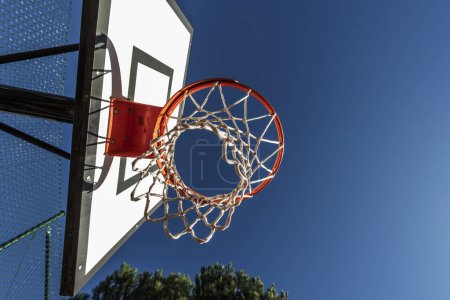 Foto de Un tiro de bajo ángulo de un aro de baloncesto con un borde naranja en una cancha al aire libre con valla metálica y árboles - Imagen libre de derechos