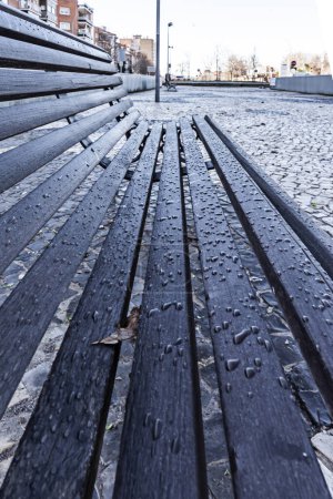 A dark wooden slatted bench filled with raindrops on an urban walkway