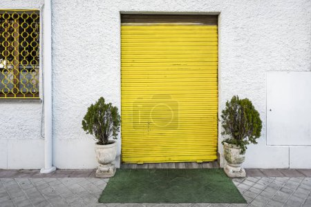 Yellow metal blinds with security closure on a white cement facade in a premises on the ground floor with some plants on the sides