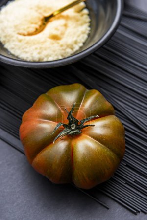 A succulent ripe raf tomato on a pile of black pasta and a bowl of grated cheese