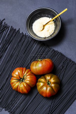 Ein Trio saftige reife Raf-Tomaten auf schwarz gefärbten Spaghetti, neben einer schwarzen Schüssel mit geriebenem Käse und einem goldenen Löffel