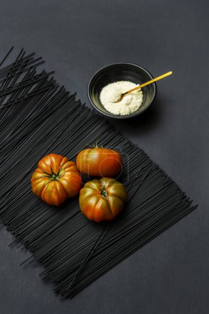 Some ripe tomatoes on a bunch of black pasta next to a black bowl with grated cheese