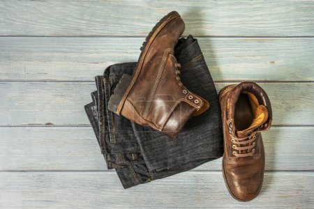 Une paire de bottes de randonnée en cuir marron avec triple couture et lacets en plastique avec jean plié sur une surface en bois clair
