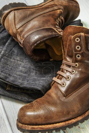 A pair of hiking boots with multiple stitching in brown leather along with folded jeans on a light wooden surface