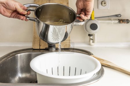 Una persona vierte agua de una olla en un colador para pasta recién hervida en un fregadero de cocina con una encimera blanca