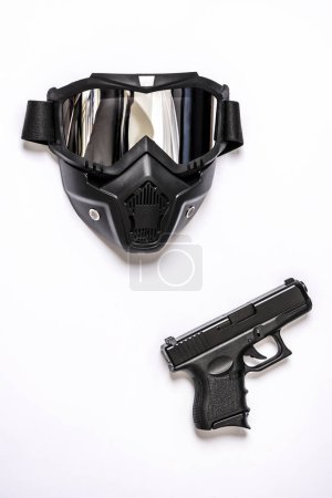 A black airsoft mask along with a mantle gun on a smooth white surface