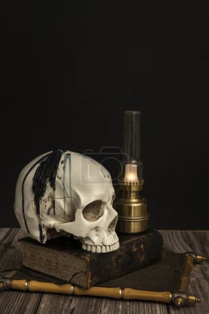 une nature morte avec un crâne blanc avec des dents supérieures sur un vieux livre à côté d'une petite lampe Argand sur une table en bois
