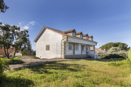Téléchargez les photos : Une maison de campagne avec des façades blanches, des sols en pierre sur le terrain, des jardins avec de l'herbe mal coupée et des arbres bas - en image libre de droit