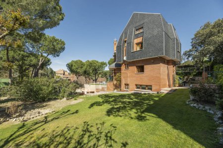 Téléchargez les photos : Une maison de campagne de plusieurs étages avec façades en brique, toits en ardoise noire, arbres et un jardin d'herbe - en image libre de droit