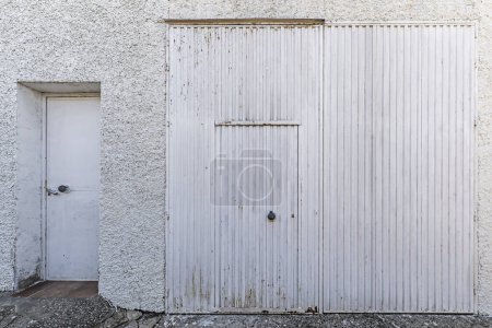 Un mur de ciment blanc rugueux avec une porte métallique blanche et une porte accessoire à l'autre