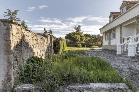Téléchargez les photos : Jardins d'une maison individuelle avec pelouse couverte, clôture en pierre avec portail métallique, façade en pierre et une petite forêt en arrière-plan - en image libre de droit