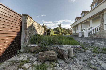 Téléchargez les photos : Jardins d'une maison unifamiliale avec pelouse envahie, clôture en pierre avec portail métallique et façade en pierre - en image libre de droit