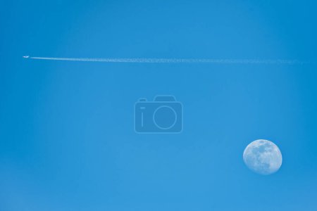 A nice picture of the moon at five in the afternoon with a passenger plane leaving a trail in the sky.