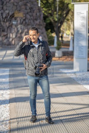 Téléchargez les photos : Jeune homme latino parlant sur un téléphone portable avec un appareil photo autour de son cou. - en image libre de droit