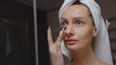Téléchargez les photos : Femme avec une serviette enroulée autour de sa tête massant son visage devant le miroir de bain. routine quotidienne du soir nettoyage du visage, soins de la peau, peeling concept de traitement de beauté - en image libre de droit