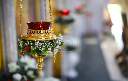 Verziert mit einem Blumenkranz, einer Lampe vor der Ikone. Eine brennende Lampe, eine Kerze in einer roten Glühbirne schwingt vor einer Ikonostase, in einem schönen Kerzenständer in einer orthodoxen Kirche. das Konzept der Orthodoxie
