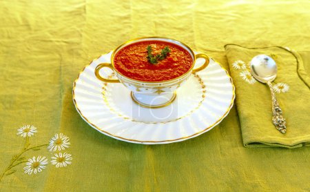 Tomato soup in a white and gold bowl on an embroidered green placemat. 