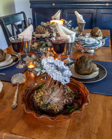 Holiday table with a rack of lamb, steamed artichoke, potatoes and on a bone China plate with candles and holiday decor.