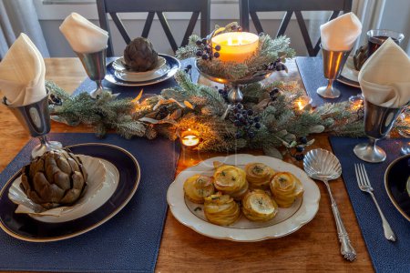 Thin sliced parmesan potatoes on a holiday table with steamed artichoke on a bone China plate with candles and holiday decor.