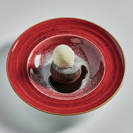 Ein Dessert, ein Schokoladen-Lavakuchen mit einer Kugel Vanilleeis, garniert mit einem Minzblatt, einer essbaren Blume und einer Physalis-Frucht, serviert auf einem leuchtend roten Keramikteller.