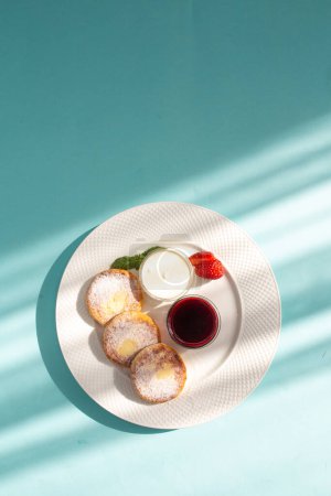 Cuatro tortitas de queso con crema agria y mermelada de fresa en un plato blanco sobre un fondo azul. Un delicioso y saludable desayuno o postre. Perfecto para un blog de alimentos o sitio web de recetas.