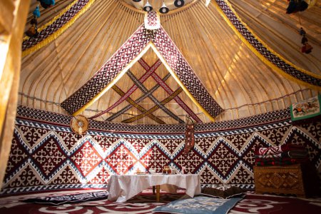 Innenraum einer zentralasiatischen Jurte mit Filzteppichen, Möbeln und einem Tisch. Traditionelle nomadische Behausung mit weißer Tischdecke.