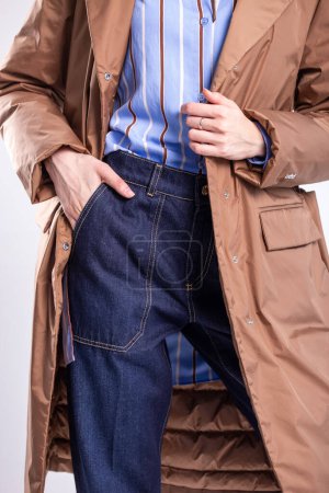Young woman in high-waisted jeans, blue shirt, brown trench coat, and nude heels posing against a white background.