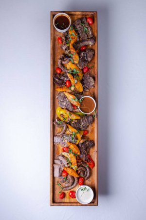 Photo for A long wooden platter filled with an assortment of meats and vegetables, including steak, sausage, chicken, potatoes, carrots, and peppers. - Royalty Free Image