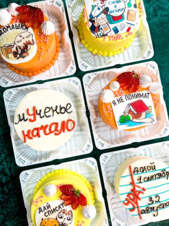 An assortment of six small square cakes with unique icing designs and decorations in plastic containers on a green surface, featuring cartoon characters, strawberries, and other decorative elements.