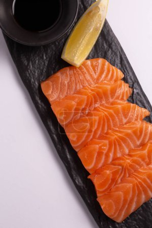 A close-up of a fresh salmon fillet garnished with lemon wedges and soy sauce on a black stone plate. This appetizing seafood dish is isolated on a white background, perfect for a healthy meal.