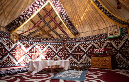 Innenraum einer zentralasiatischen Jurte mit Filzteppichen, Möbeln und einem Tisch. Traditionelle nomadische Behausung mit weißer Tischdecke.