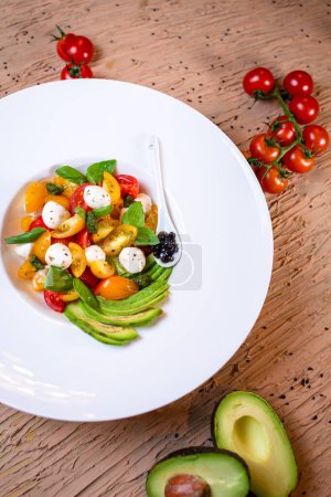 Healthy salad with tomatoes, avocado, mozzarella, basil on white plate. Garnished with balsamic glaze, served with bread.